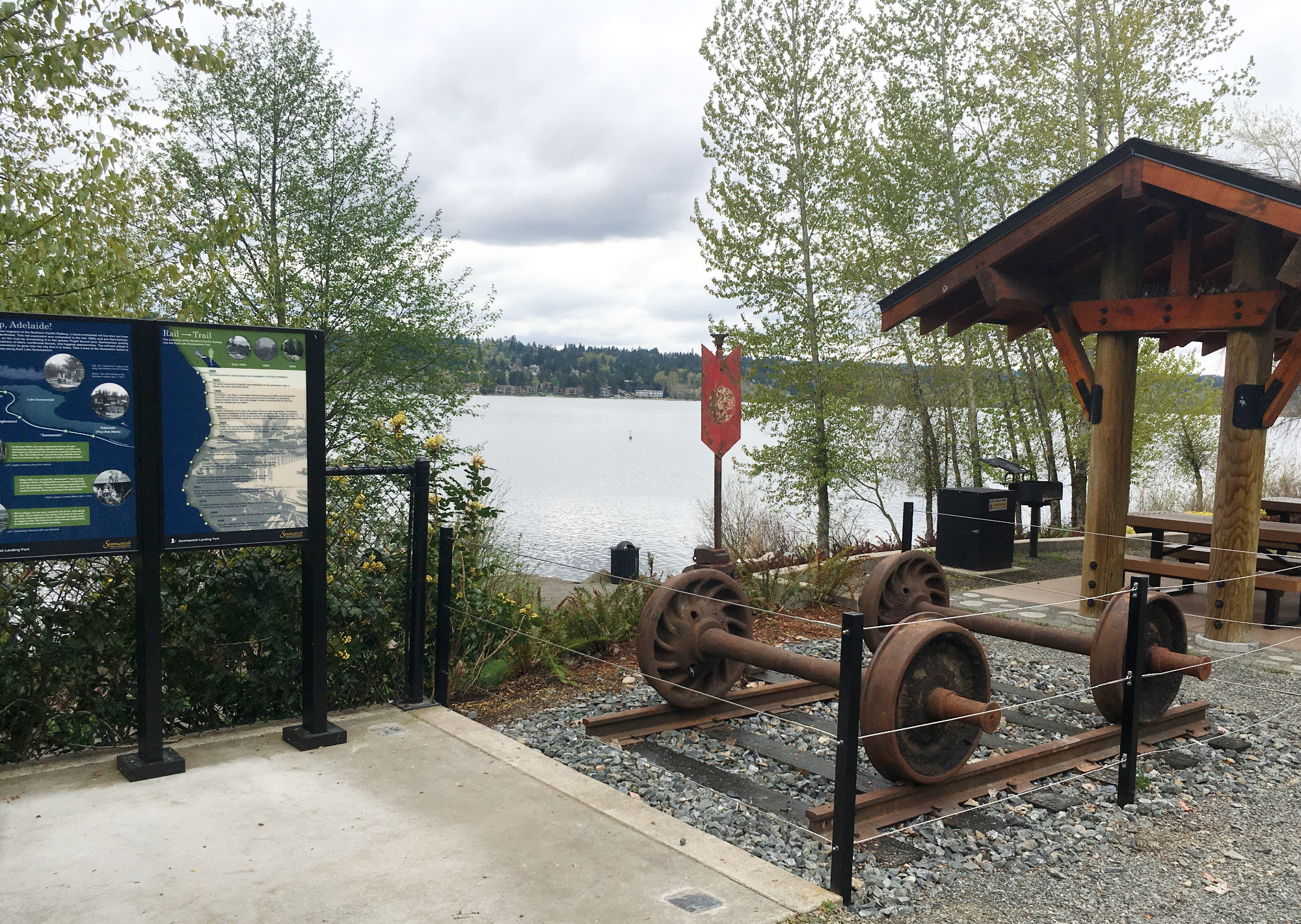 Antique train axles are displayed at Sammamish Landing Park next to educational signage, a picnic shelter, and Lake Sammamish.