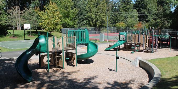Two playground units with slides, climbing bars, and other attachments at Northeast Sammamish Park. A tennis court and basketball court are in the background.