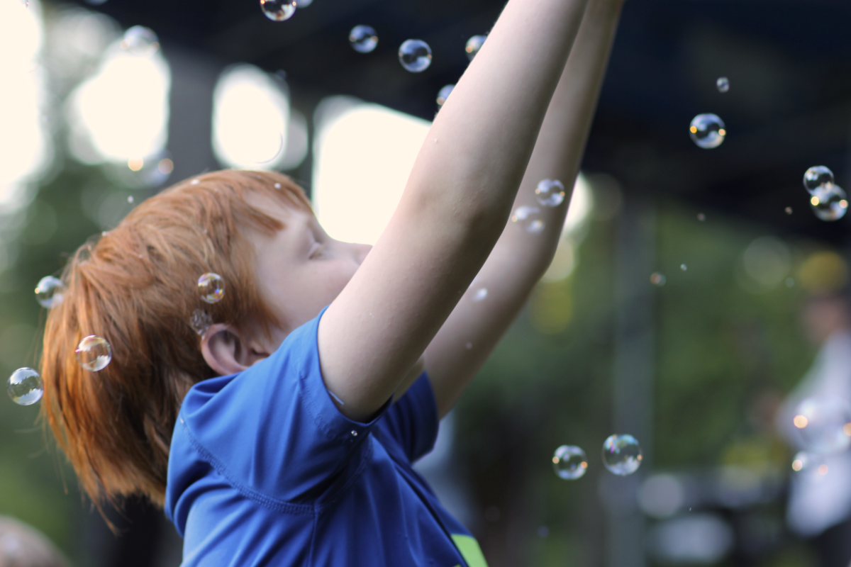 Child with red hair and blue shirt dancing with bubbles at a Concert in the Park