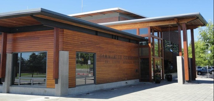 A modern building with wood and glass exterior, overhanging roof, and large windows, surrounded by trees and parked cars under a clear blue sky at Sammamish City Hall.