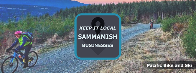 Two cyclists pedaling on gravel trail with trees in the background and a valley and mountain range with an orange sky off to the right. The message in the center reads Keep it Local Sammamish Businesses. The bottom right says Pacific Bike and Ski.
