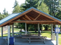 East Sammamish Park picnic shelter with blue metal roof and four picnic tables. There are four garbage/recycle bins. The shelter is surrounded by a grassy area, trees, and near a fenced field.