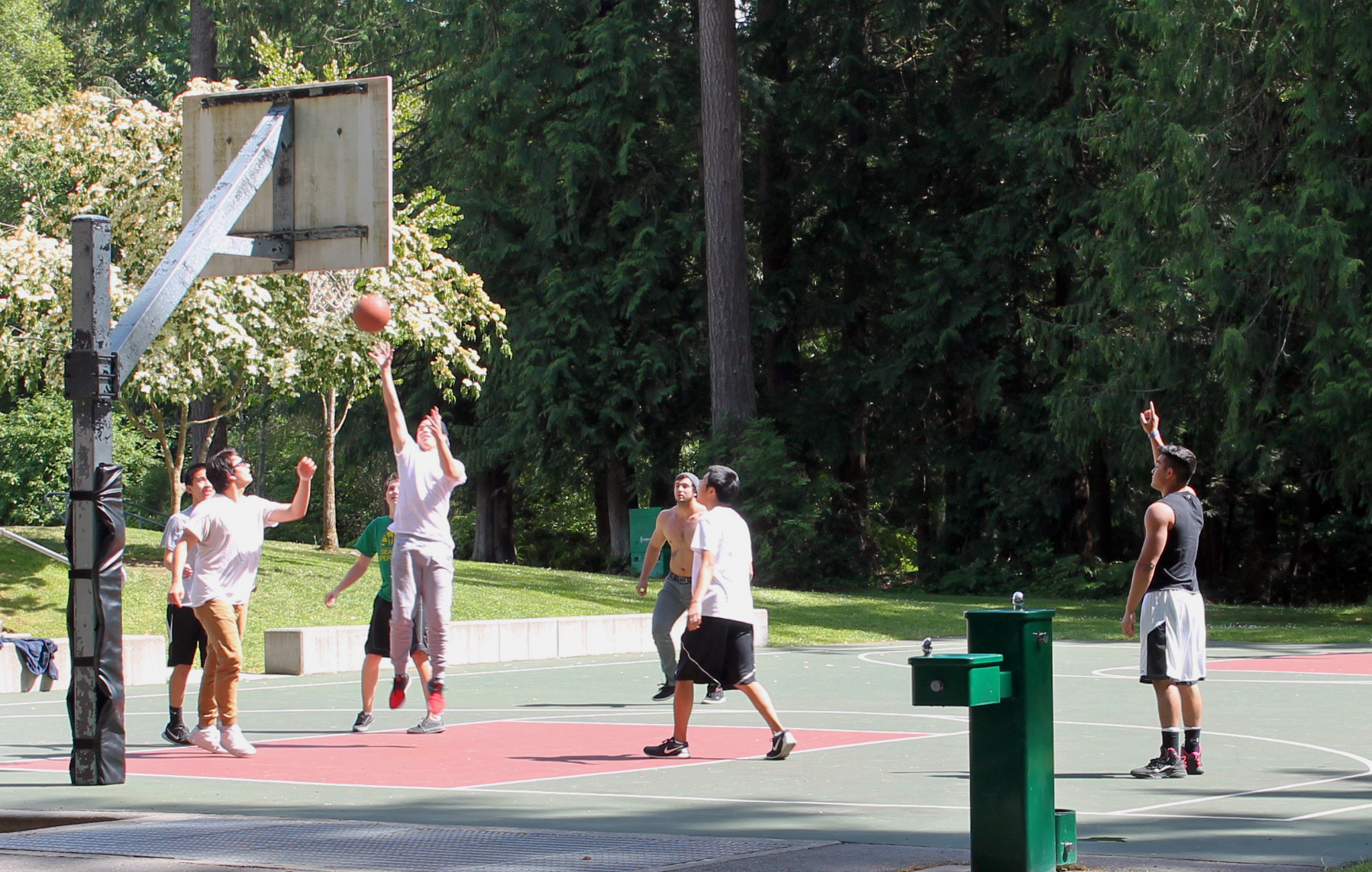 A group of young people in street wear plays basketball at pine lake park on a sunny day. There is an accessible drinking fountain next to the court.