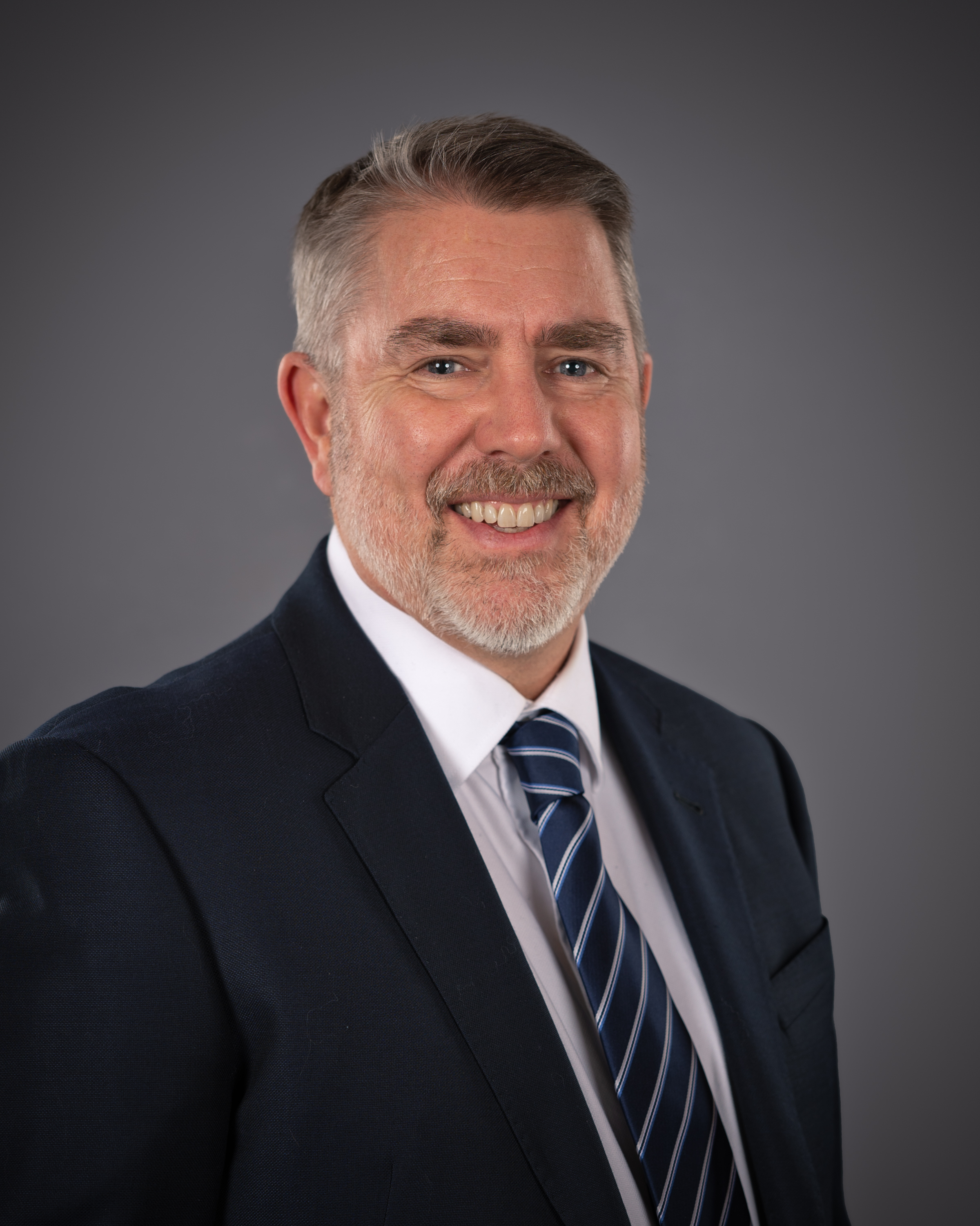 A middle-aged man with short gray hair and a beard, wearing a navy suit, white shirt, and striped tie, smiles against a plain gray background.