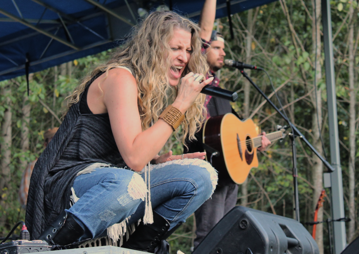 Woman in blue jeans and black tank top sings powerfully at a Sammamish Concert in the Park while a guitar player stands in the background.