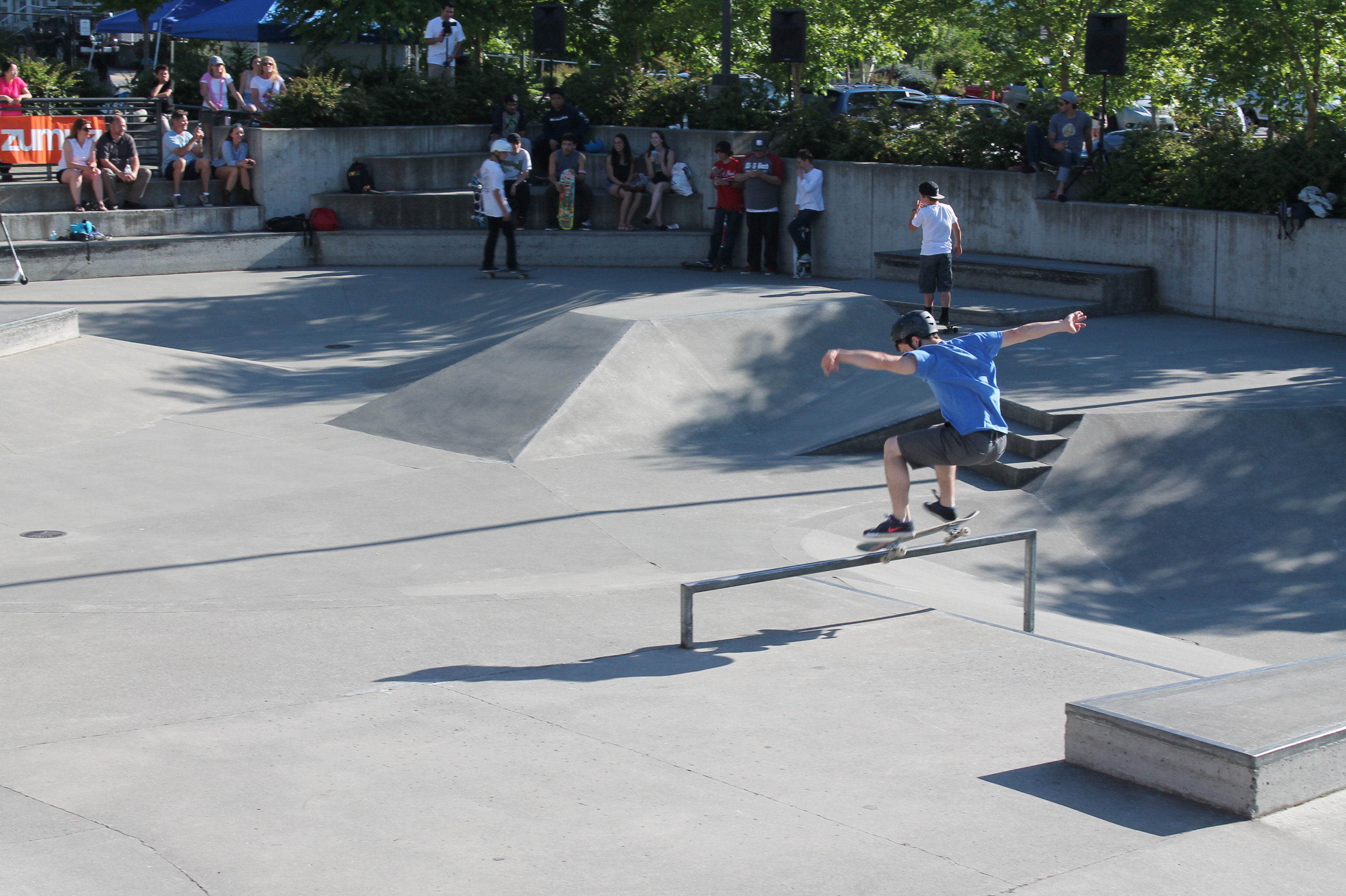 skateboarding kid in helmet does rail trick at Sammamish's skate park while onlookers watch from built-in seats and steps