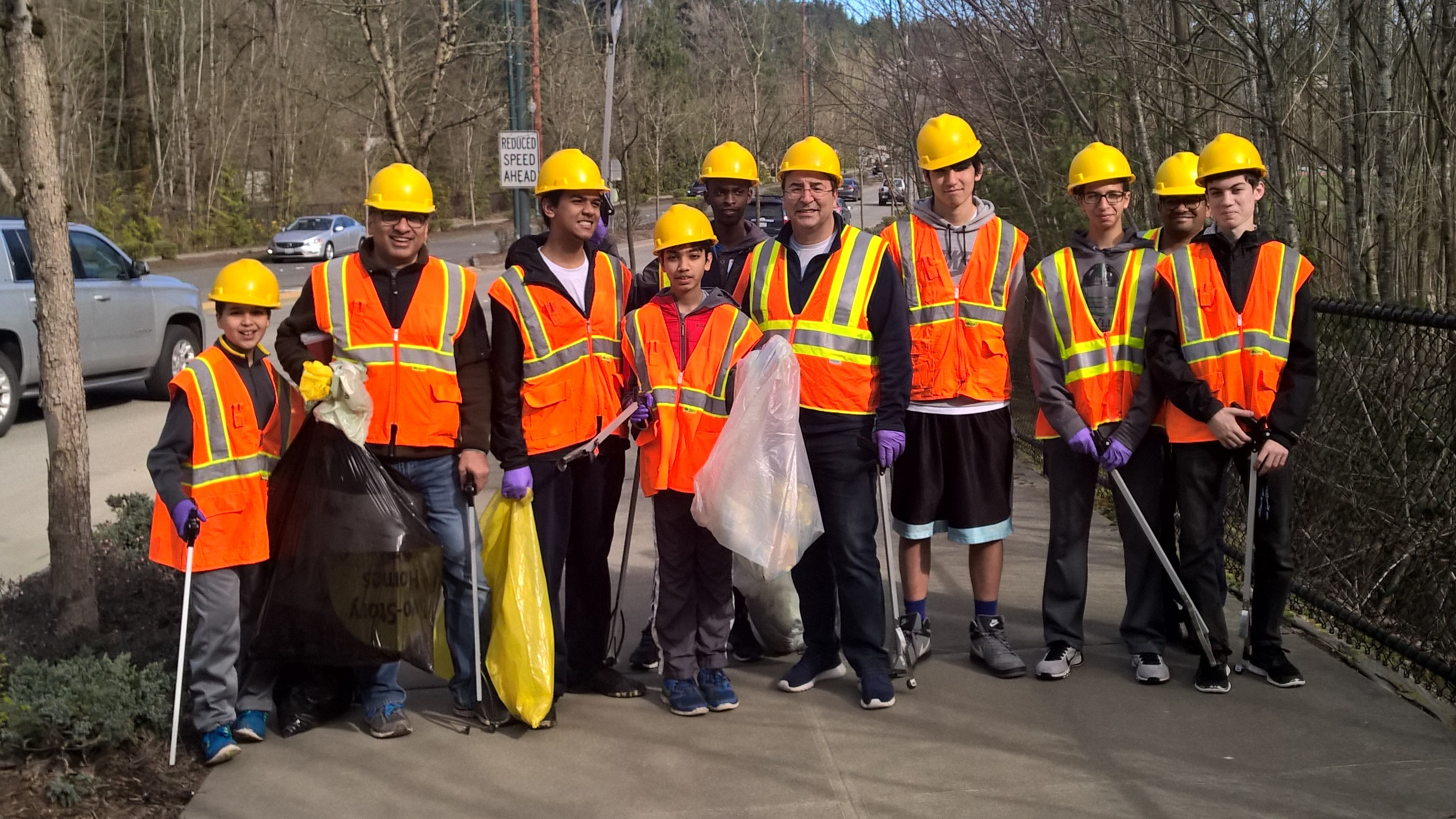 A group of ten adopt-a-road volunteers pose next to a Sammamish city street wearing bright orange construction worker vests and yellow hard hats and holding plastic garbage bags and trash grabbing gadgets. 